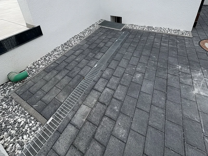 Neu gepflasterte Fläche am Haus aus dunklem Rechteckpflaster mit Entwässerungsrinne und Zierkiesstreifen